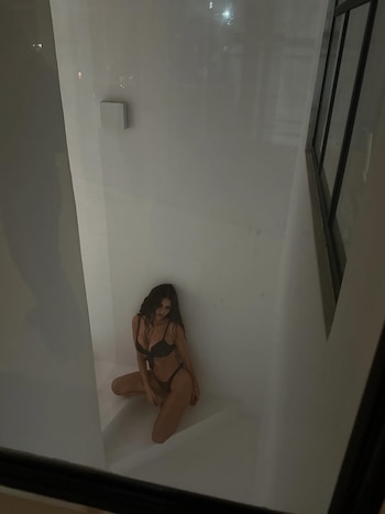 Zaira Nara, con cabello oscuro y largo, posa sentada en cuclillas vistiendo ropa interior negra sobre una superficie blanca en un espacio minimalista