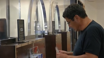 Hombre con gafas y camiseta oscura mira hacia abajo sujetando papeles en un mostrador con un monitor de computadora y paneles transparentes