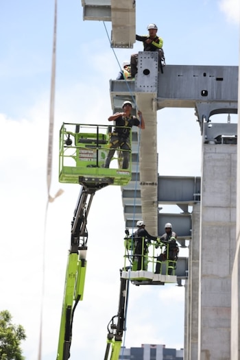 Imagen de un trabajador en una plataforma elevadora junto a grandes pilares de concreto y estructuras metálicas grises en construcción, con una grúa y cielo azul