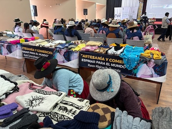 Dos mujeres artesanas sentadas en el suelo, rodeadas de coloridas prendas tejidas a mano. Al fondo, varias personas sentadas en un salón y mesas con productos textiles