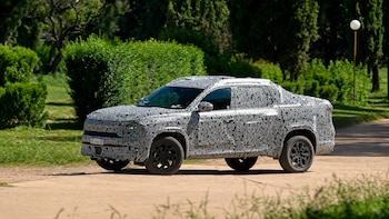 Un prototipo de pickup camuflado con un patrón blanco y negro avanza por un camino de tierra, rodeado de vegetación verde y árboles bajo un cielo soleado
