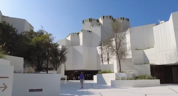 Edificio de fachada blanca con formas geométricas escalonadas, vegetación colgante y árboles. Una persona camina. Dos esculturas blancas de órix