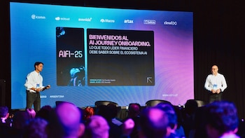 Dos hombres en un escenario. Uno habla frente a una pantalla con texto sobre IA y logos empresariales; el otro observa. Una audiencia parcialmente visible