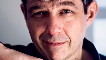Primer plano de un hombre de mediana edad con cabello oscuro y canoso, piel clara con pecas, y ojos oscuros, sonriendo; lleva camisa oscura