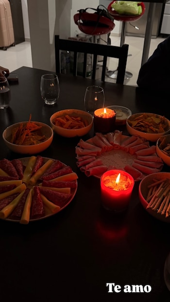 Una mesa con platters de embutidos y queso, bowls con snacks, dip, tres velas encendidas, copas de vidrio y una maleta en el fondo