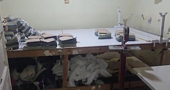 Sala de taller desordenada con una mesa de trabajo larga llena de pilas de piezas de material marrón y verde, herramientas de corte y materiales debajo de la mesa