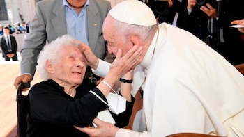 Primer plano del Papa Francisco, un hombre mayor, sentado y vestido con una sotana blanca, con la mano derecha levantada hacia la oreja, y un micrófono delante. Se ve una cruz pectoral