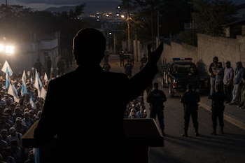 Silueta de un hombre en un atril frente a personas con banderas guatemaltecas, patrulla y agentes policiales en una calle nocturna.