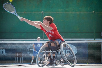 Gonzalo Lazarte, un hombre joven, juega tenis en silla de ruedas, vistiendo una camiseta roja y pantalones cortos oscuros. Alza una raqueta para golpear una pelota amarilla en una cancha