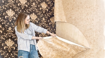 Mujer con camisa a rayas y jeans retira papel de pared beige, revelando un papel pintado oscuro con estampado floral en la pared de una habitación.