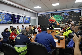 Un grupo de personas sentadas en una sala de conferencias. Varias pantallas en la pared muestran mapas geográficos y datos. Algunos llevan uniformes