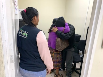 Un niño con chaqueta negra está de pie junto a un adulto con chaleco que apoya su mano en el hombro del menor en una sala