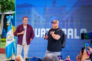 Dos hombres en un escenario con un micrófono, frente a una pantalla con texto "GUATEMALA" y arquitectura, junto a una bandera nacional. Público con teléfonos