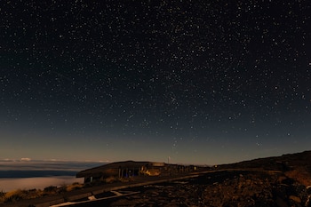 Cielo nocturno con estrellas sobre una carretera asfaltada, un edificio bajo y un mar de nubes iluminado en el horizonte