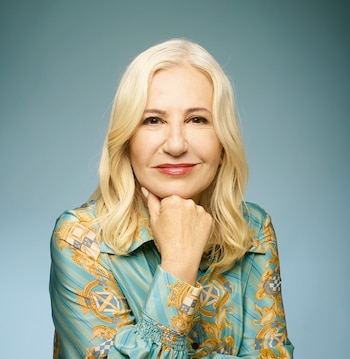 Retrato de mujer de mediana edad con cabello rubio, blusa estampada verde y dorada, sonriendo con la barbilla en la mano derecha, sobre fondo azul claro
