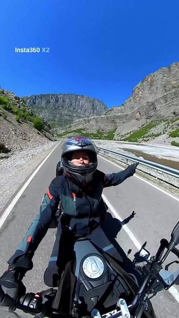 Persona con casco y traje de motociclista conduce moto por carretera montañosa. Río seco y vallas a la derecha, cielo azul despejado. Vista frontal