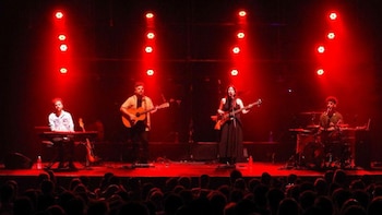 Cuatro músicos en un escenario bajo luces rojas; un hombre toca el teclado, otro la guitarra, una mujer el bajo y canta, y otro hombre la batería