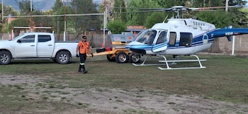 Un helicóptero blanco y azul en el césped, junto a una camioneta blanca con un remolque cisterna y un hombre con chaleco naranja