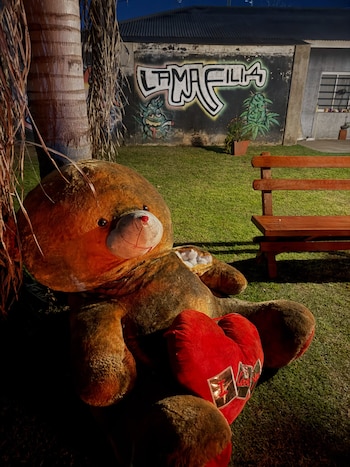 Un oso de peluche gigante con un corazón rojo descansa en el césped frente a un muro con un grafiti de La Mafilia y un banco de madera