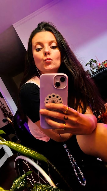 Una mujer con cabello oscuro y blusa negra se toma un selfie con un teléfono morado, haciendo una boca de pato. Parte de una silla de ruedas es visible