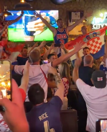 Multitud animando y celebrando en un bar. Un hombre con camiseta de baloncesto levanta los brazos. Se ven banderas croatas, una pantalla y teléfonos móviles