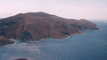 Vista aérea de una gran montaña árida con vegetación escasa que desciende abruptamente hacia un mar azul brillante, con un pequeño bote visible cerca de la costa