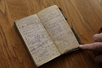 Libro de contabilidad antiguo abierto sobre madera. Muestra páginas con registros manuscritos en tinta violeta. Un dedo señala la página derecha