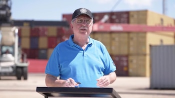 Un hombre de mediana edad con gorra negra, gafas y polo azul claro habla al aire libre, con contenedores de carga y una grúa al fondo