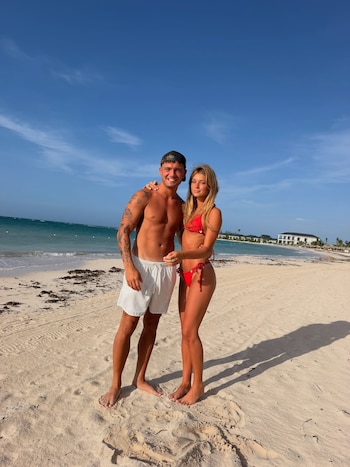 Hombre con shorts blancos y tatuajes junto a mujer en bikini rojo, de pie en una playa. Algas, mar, cielo azul y edificios blancos distantes