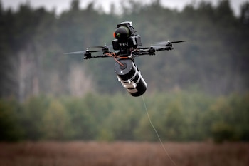Un dron negro con cuatro hélices y un dispositivo cilíndrico del que cuelga un delgado cable, vuela contra un fondo borroso de árboles