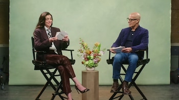 Anne Hathaway y Stanley Tucci sentados en sillas negras de director, sosteniendo tarjetas de "VOGUE", frente a un fondo verde claro con un arreglo floral