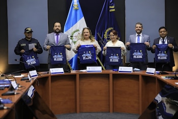Seis personas sostienen bolsas azules con texto impreso detrás de una mesa de reuniones frente a la bandera de Guatemala