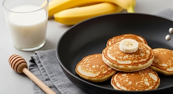 Panqueques de banana dorados en sartén negra, con rodaja de plátano, un vaso de leche, bananas enteras y un dosificador de miel sobre mesa gris.