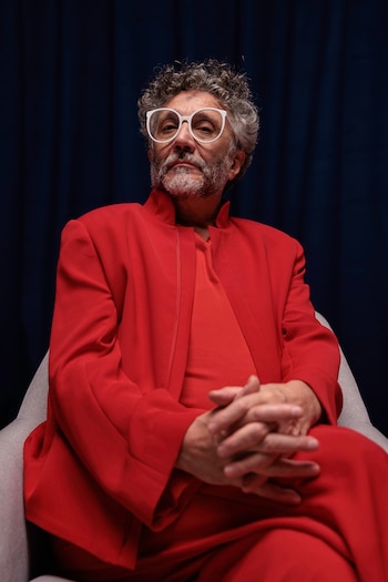 Retrato de Fito Páez sentado en una silla gris, vistiendo un traje rojo y gafas blancas, con el pelo y la barba canosos. Fondo azul oscuro