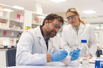 Un hombre y una mujer con batas blancas y gafas protectoras manipulan recipientes e instrumentos sobre una mesa de laboratorio