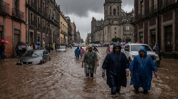 Calle urbana de México con agua marrón cubriendo la calzada hasta los neumáticos de los coches. Personas con impermeables caminan y un edificio histórico se ve al fondo.