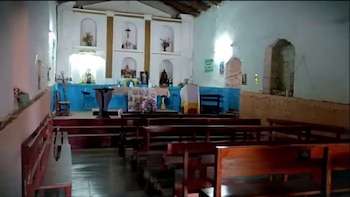 Interior de una iglesia con bancas de madera oscura y paredes blancas. Se ve un altar con un paño azul, figuras religiosas en nichos y un crucifijo en la pared