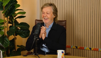 Paul McCartney sentado frente a un micrófono y una taza sobre una mesa, con una planta y un panel de madera detrás