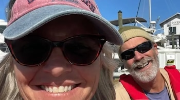 Un hombre y una mujer sonrientes, con gafas de sol, en un barco. Él lleva gorro y chaleco salvavidas. Ella, visera. Embarcaciones al fondo