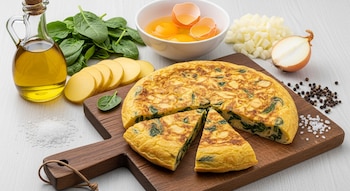 Tortilla de papas y espinaca con dos porciones cortadas sobre una tabla de madera, rodeada de ingredientes como aceite de oliva, espinacas, patatas, huevos, cebolla, sal y pimienta.