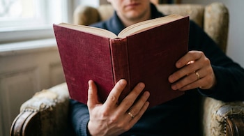 Primer plano de las manos de una persona sosteniendo un libro abierto de tapa dura bordó, sentada en un sillón, con el rostro parcialmente visible al fondo.