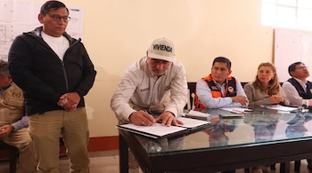 Un hombre con gorra firma un documento en una mesa. Otro hombre de pie y tres personas sentadas observan. Un plano técnico está colgado en la pared