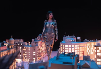 Una mujer con vestido plateado se erige sobre una maqueta de ciudad iluminada con edificios, calles, automóviles y un fondo negro