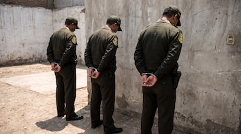 Tres hombres de espaldas con uniforme de policía verde oscuro y gorra verde, esposados, parados frente a una pared de concreto agrietada.