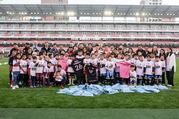 Gran grupo de niños y adultos posando en un campo de fútbol con gradas de estadio rojas al fondo. Los niños visten camisetas rosas y algunos adultos sostienen camisetas deportivas