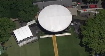 Vista aérea de una gran carpa blanca circular y una carpa blanca rectangular adyacente sobre césped verde. Hay árboles alrededor y algunos edificios y vehículos pequeños