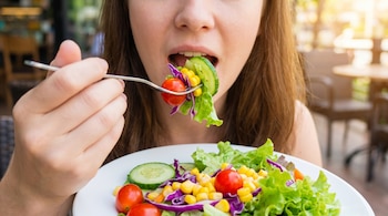 Primer plano de una persona comiendo una ensalada colorida con lechuga, pepino, tomate cherry, maíz y col morada. Se ve un tenedor y parte de un plato blanco.