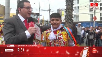 Un reportero con micrófono entrevista a un joven bailarín vestido con traje típico de la danza de las tijeras, en una calle con público y edificios