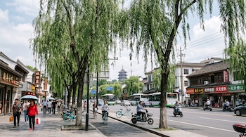 Árboles de sauce en una acera junto a una calle con peatones, ciclistas, motocicletas, autobuses y comercios con carteles en chino.