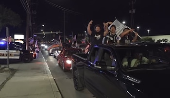 Vista nocturna de un desfile de vehículos; varios aficionados, incluyendo uno con camiseta de los Spurs, celebran de pie en camionetas y coches